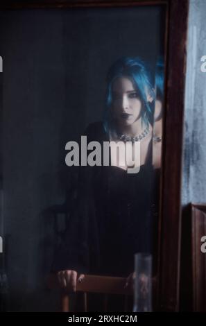 Indoors portrait of gorgeous goth girl in black dress. Blue-haired sad gothic lady looking into the old dirty mirror. Vintage style Stock Photo