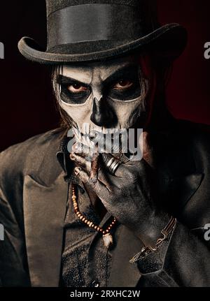 A young man in image of Baron Samedi, the Voodoo deity. Baron Saturday dressed in black suit and top hat smoking cigar, close-up portrait. Day of the Stock Photo