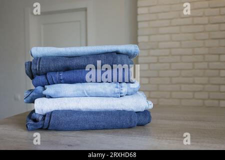 A neatly folded stack of clothes - jeans in different shades of blue on a wooden table against the backdrop of a bright room. Stock Photo
