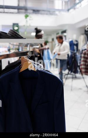 Formal male blazers hanging on rack in fashion boutique with blurred background. Male jackets latest arrival on hangers and accessories on shelf in department shopping mall close up selective focus Stock Photo