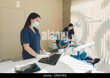 Female dentist typing patient data on laptop computer while patient converses with assistant in dental office chair. Stock Photo