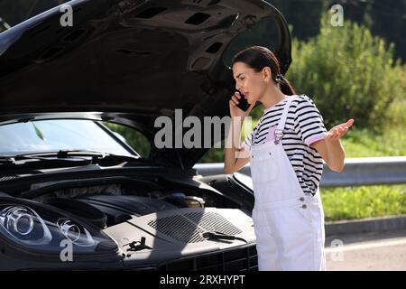 Stressed woman talking on smartphone while looking under hood of broken car outdoors Stock Photo