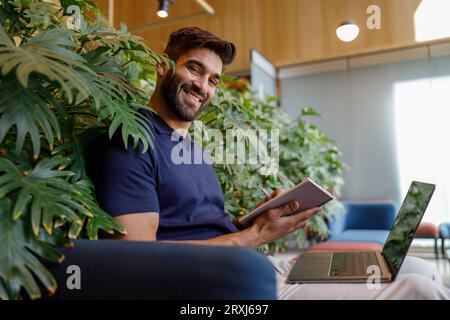 Handsome smiling businessman is making notes during work on laptop in coworking and looks camera Stock Photo