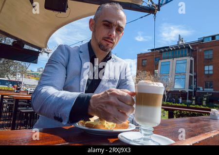 Caucasian business adult man in elegant clothes, taking cup of cappuccino coffee from counter, sitting having breakfast outside restaurant. Stock Photo