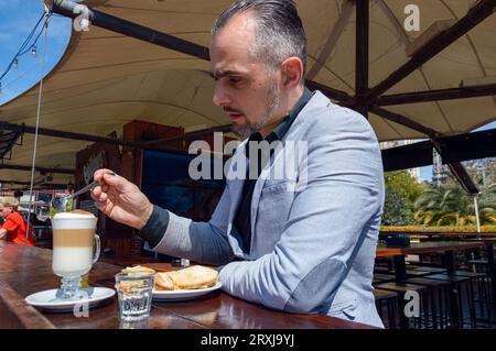 Caucasian adult man of French ethnicity wearing elegant clothes, sitting having breakfast having coffee with sandwich sitting at bar outside restauran Stock Photo