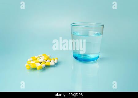 white and yellow medical pills, vitamins, in the distance there is a beautiful blue glass with clean water, on a harmonious bright blue background. Ta Stock Photo