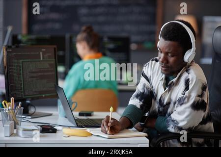 Serious programmer in headphones making notes in notebook while working with security codes on computer at his workplace Stock Photo