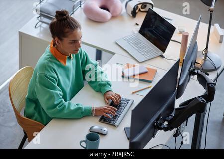 High angle view of young woman sitting at table in front of computer monitor and typing Stock Photo