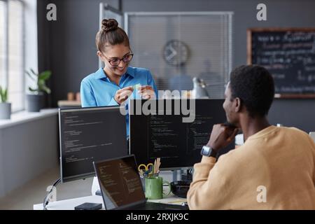 Young woman talking to her colleague while he writing codes on computer at his workplace Stock Photo