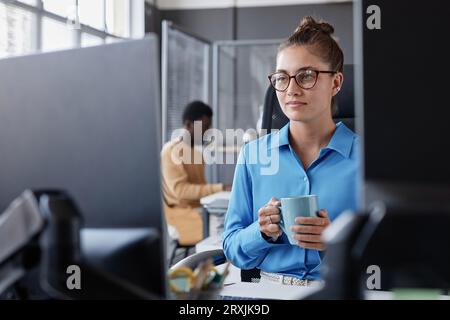 Young woman sitting at her workplace and looking at computer monitor while drinking coffee Stock Photo