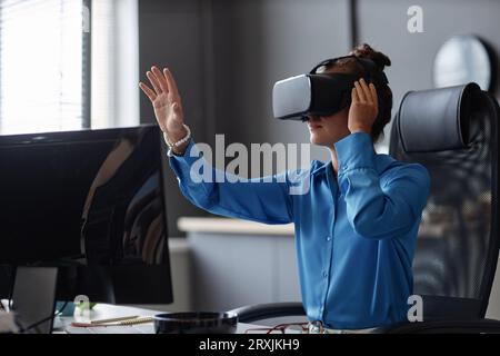 Young woman testing new program in VR glasses while sitting at her workplace Stock Photo