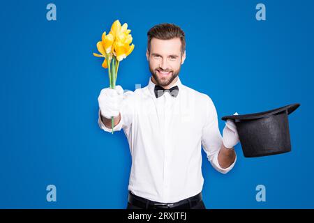 Portrait of romantic positive magician with bristle and modern hairstyle getting a yellow flowers from tophat and giving it front, isolated on grey Stock Photo