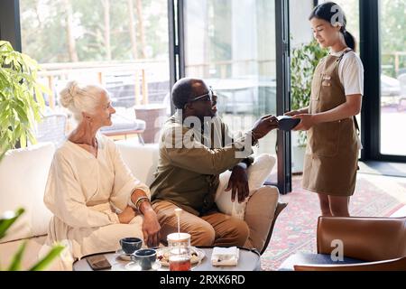 Multiethnic senior couple paying for coffee while sitting in cafe Stock Photo