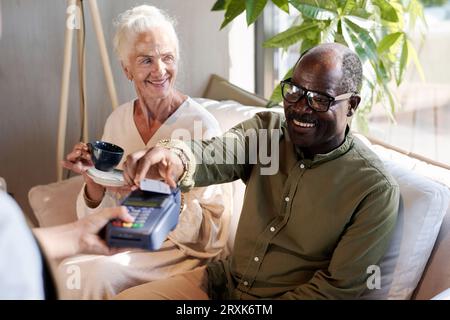 Smiling African American man paying for coffee with credit card while sitting in cafe with his wife Stock Photo