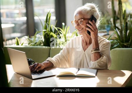 Smiling senior woman talking on mobile phone and typing on laptop while sitting at table in cafe Stock Photo