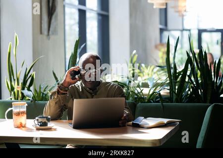 Senior man using laptop and talking on the phone while sitting in cafe Stock Photo