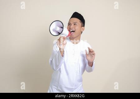 Portrait of attractive Asian muslim man in white shirt with skullcap speaking louder using megaphone, promoting product. Advertising concept. Isolated Stock Photo