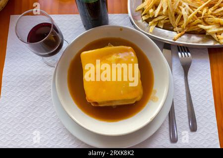 Traditional portuguese sandwich francesinha with glass of wine and fried potato. Francesinha in hot sauce with fork and knife. Portuguese cuisine. Stock Photo