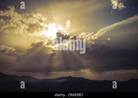Golden sunset behind clouds whit sun rays through the silhouetted clouds.  the mountains its backgroand in summer day.. Stock Photo