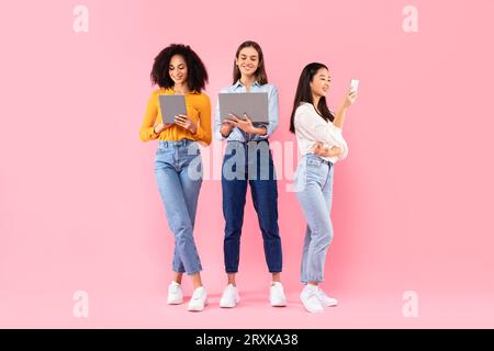 Happy multiracial ladies friends using different gadgets, using smartphone, laptop and digital tablet, pink background Stock Photo