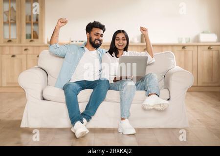 Giveaway, lottery, cashback. Overjoyed indian couple using laptop sitting on couch at home, gesturing and exclaiming Stock Photo