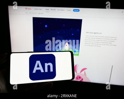 Person holding cellphone with logo of computer animation software Adobe Animate on screen in front of company webpage. Focus on phone display. Stock Photo