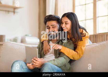 Technologies for leisure. Indian couple using digital tablet, having video call with friends, resting on sofa at home Stock Photo