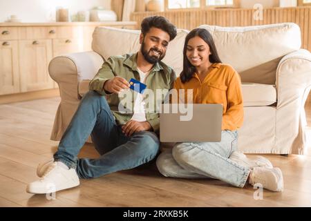 Happy indian spouses shopping online via laptop and using credit card, sitting on floor at home interior, copy space Stock Photo