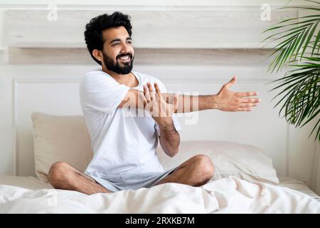 Young Indian Man Stretching Arm Muscles While Sitting In Bed After Awake Stock Photo