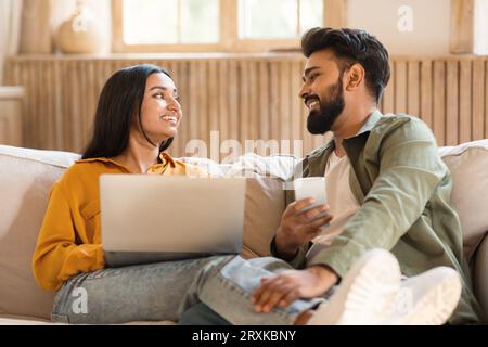 Weekend pastimes concept. Happy indian couple using laptop and smartphone, looking at each other while relaxing on couch Stock Photo