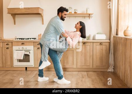 Romantic indian couple having fun together, feeling happy, dancing in kitchen interior, full length, side view Stock Photo