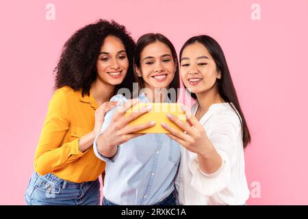 Happy multiracial female friends making selfie on cellphone, taking photos for social media over pink background Stock Photo
