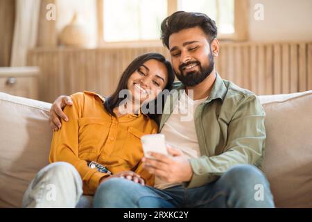 Happy loving hindu couple using cellphone, having video call or browsing internet, man embracing wife, sitting on sofa Stock Photo