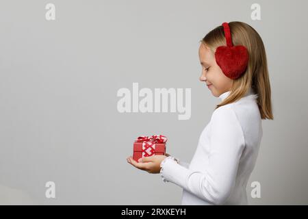 Cute little girl wearing red earmuffs holding gift present box on white studio wall background. Child, profile portrait Stock Photo