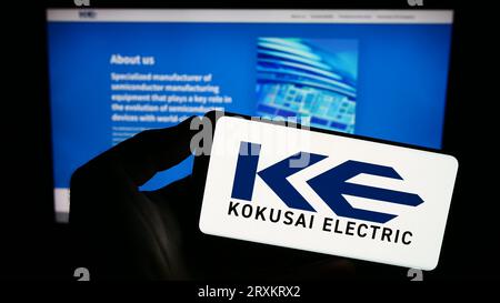 Person holding smartphone with logo of Japanese company Kokusai Electric Corporation on screen in front of website. Focus on phone display. Stock Photo