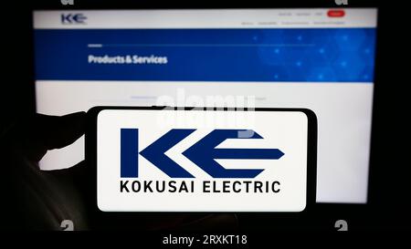 Person holding mobile phone with logo of Japanese company Kokusai Electric Corporation on screen in front of web page. Focus on phone display. Stock Photo