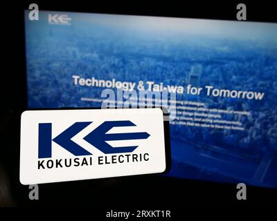 Person holding cellphone with logo of Japanese company Kokusai Electric Corporation on screen in front of webpage. Focus on phone display. Stock Photo