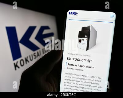 Person holding cellphone with webpage of Japanese company Kokusai Electric Corporation on screen with logo. Focus on center of phone display. Stock Photo