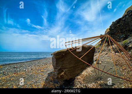 Rusty beach parasol carcass resting on rock, black and grey volcanic pebble and sand beach, blue sky and clouds, calm sea tide, rocky cliffs, sunny day. Santorini island, Greece.  Stock Photo