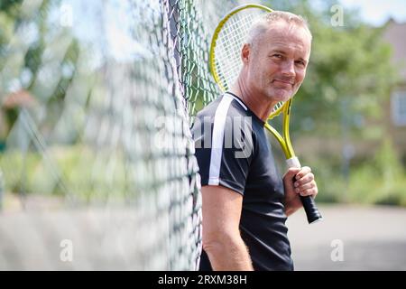 Mature man holding tennis racket leaning on fence Stock Photo