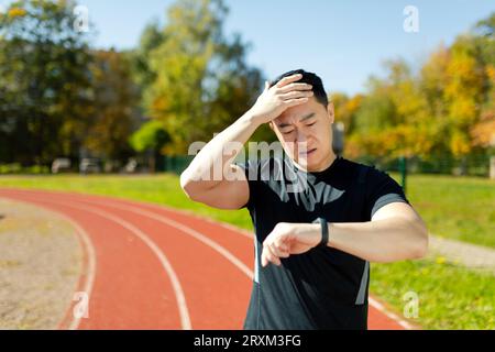 Asian young male athlete and runner standing in stadium, holding head and looking upset at tracker, unhappy and disappointed with result. Stock Photo