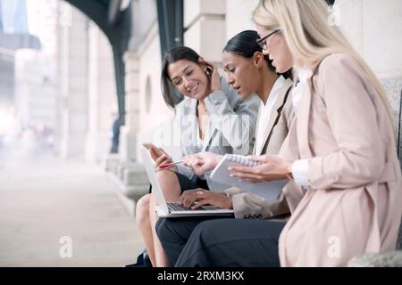 Colleagues using smart phone and laptop on bench Stock Photo