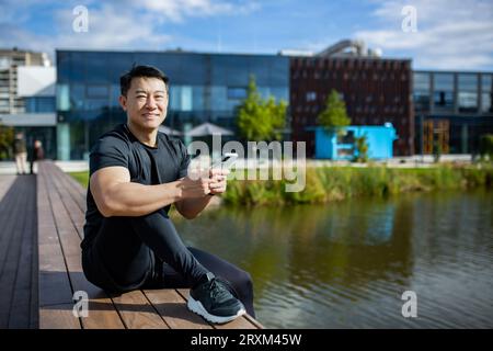 Portrait of an Asian young man in a black sports jacket sitting on a bench by the lake in the park, using a mobile phone and resting. Smiling at the camera. Stock Photo
