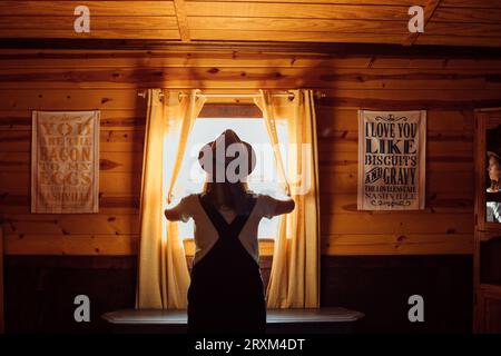 Woman wearing hat opening curtains in wooden cabin Stock Photo