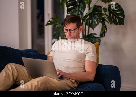 Man middle aged student working on educational project lying on couch at home in evening after job. Stock Photo