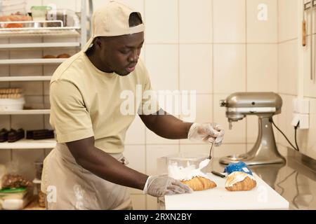 Side view portrait of young male baker decorating fresh croissants with cream in artisan bakery, copy space Stock Photo