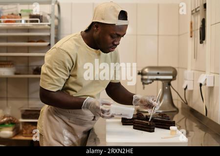 Side view portrait of young Black man as male baker decorating chocolate pastries in bakery, copy space Stock Photo