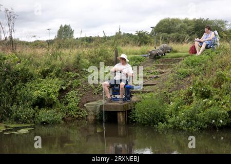 Man sitting on raised platform fishing with rod and line in River Avonwith wife sitting patiently readibng near Evesham UK Stock Photo