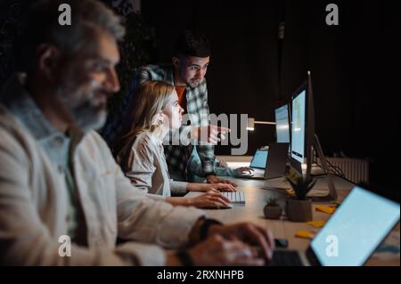 Focused caucasian guy wearing plaid shirt typing pc computer in cozy evening office with his colleagues Stock Photo