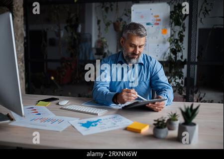 Senior handsome man sitting at the table in office, working from home on tablet computer Stock Photo